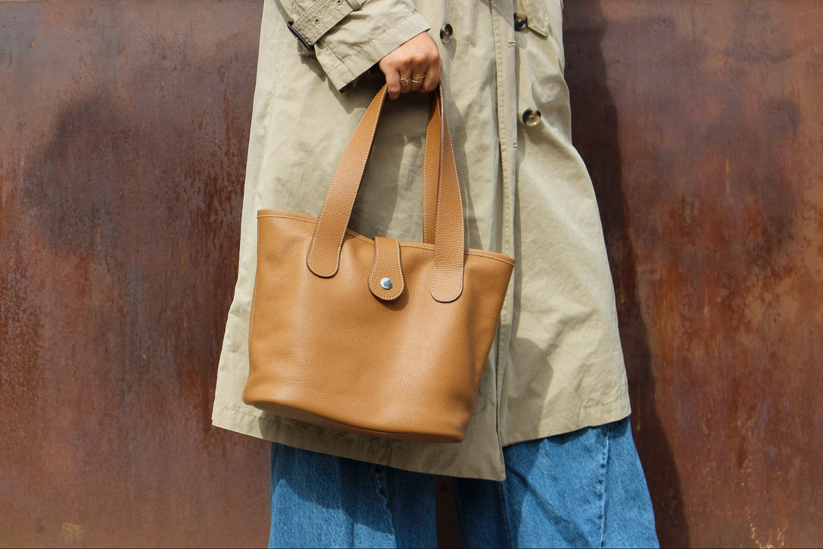 Sac cabas Saint Barth en cuir grainé camel avec patte et bouton pression argenté, porté à la main avec un trench beige et un jean.